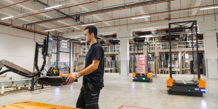 Person operating machinery in a warehouse, holding a control device while monitoring equipment. Automated guided vehicles (AGVs) transport goods in the background. The warehouse features high ceilings with exposed pipes and lighting.