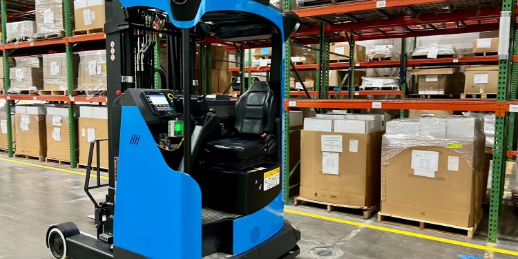 Blue and black forklift in a warehouse, positioned in front of multi-level shelving filled with large boxes. Yellow floor markings indicate pathways and storage zones.