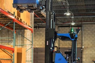 Automated blue and black forklift lifting a large blue container in a warehouse with high ceilings, shelving, and visible safety signage.