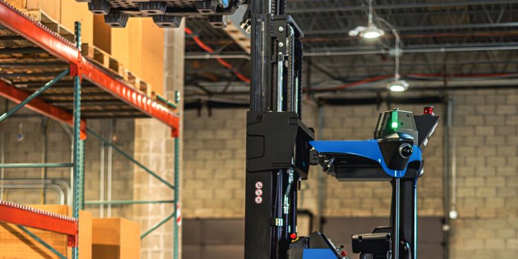 Automated blue and black forklift lifting a large blue container in a warehouse with high ceilings, shelving, and visible safety signage.